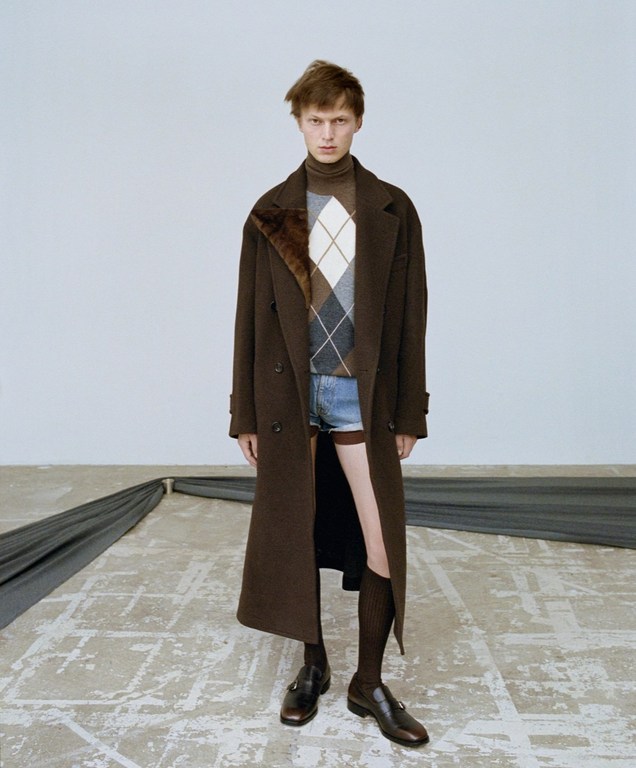 Fashion portrait of a young adult male wearing a brown coat, argyle sweater, and jean shorts standing in a studio with dark brown shoes.