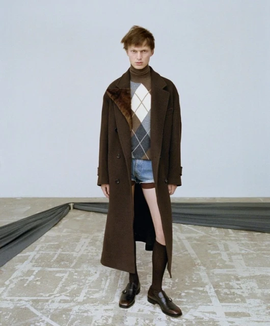 Fashion portrait of a young adult male wearing a brown coat, argyle sweater, and jean shorts standing in a studio with dark brown shoes.