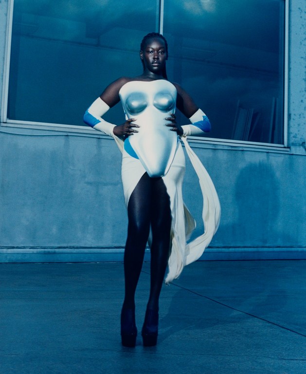 An adult female with dark skin poses in a futuristic white dress with silver accents and blue details in a blue-toned outdoor setting.