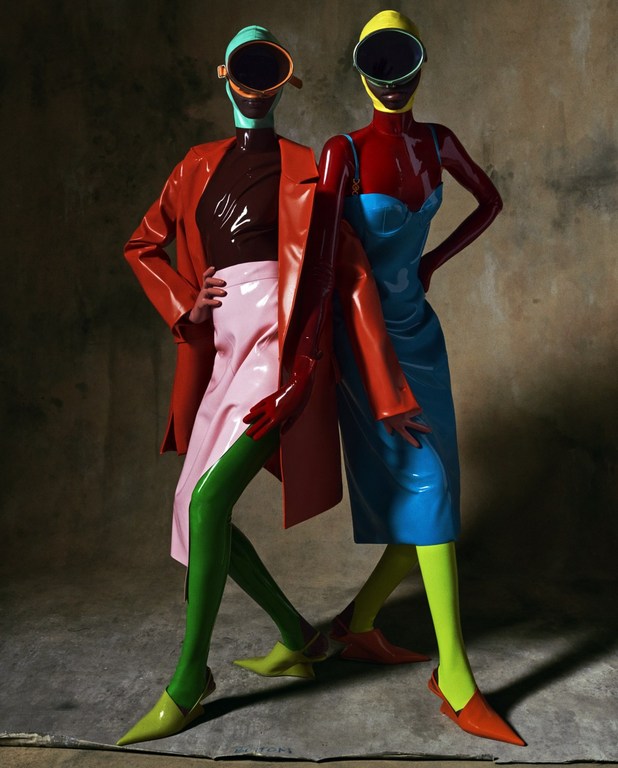 Two models in matching latex outfits pose in front of a mottled backdrop, wearing helmets with visors and colorful dresses and accessories.