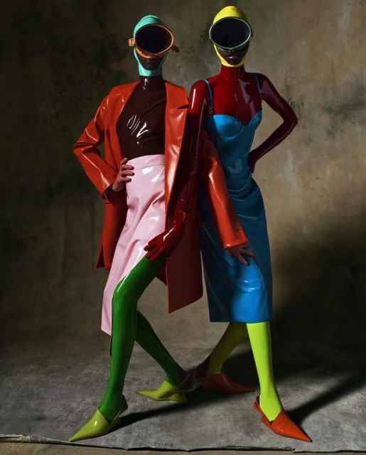 Two models in matching latex outfits pose in front of a mottled backdrop, wearing helmets with visors and colorful dresses and accessories.