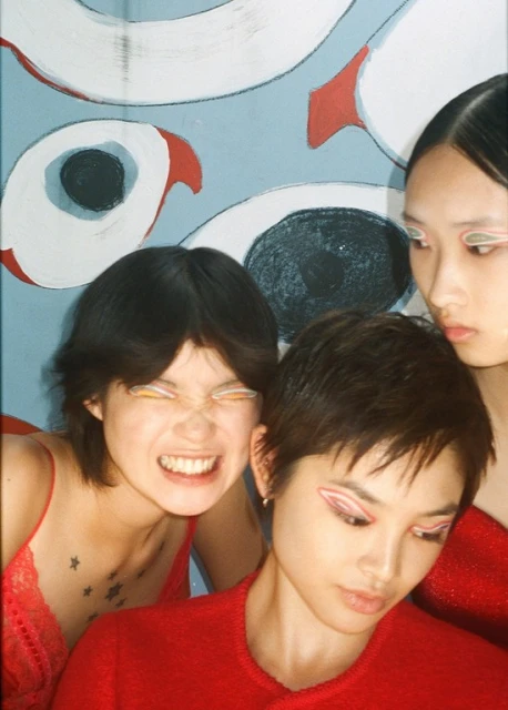 Three young women pose in a photo studio with theatrical eye makeup and red outfits, standing in front of an abstract painted backdrop.