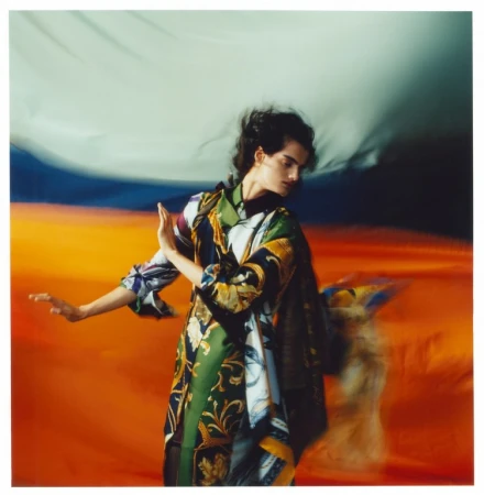 Model in patterned silk garment posing against a vibrant abstract orange and blue backdrop