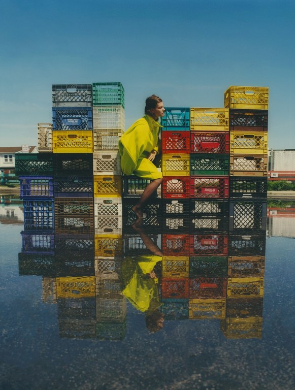 Woman in yellow dress posing on a colorful milk crate wall reflected in water, clear blue sky in the background on a sunny day.
