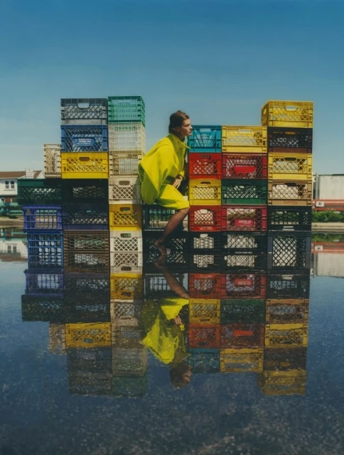 Woman in yellow dress posing on a colorful milk crate wall reflected in water, clear blue sky in the background on a sunny day.