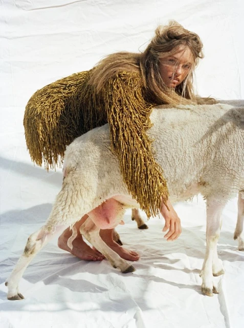 Woman in fringed coat leans on a sheep in a bright studio setting, showcasing fashion and nature with a surreal, artistic composition.