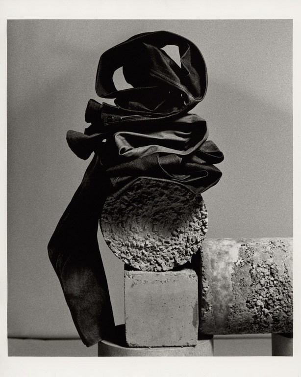 Black and white still life photograph of folded trousers stacked atop textured concrete cylinders and blocks.