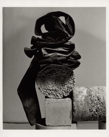 Black and white still life photograph of folded trousers stacked atop textured concrete cylinders and blocks.
