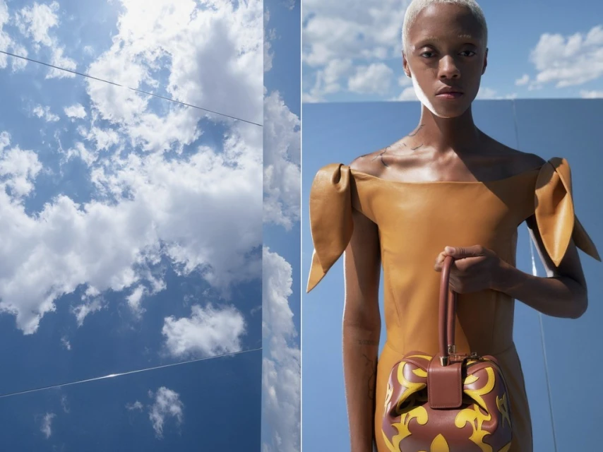 Blonde model wearing a tan leather dress and holding a brown patterned handbag, posing outdoors with a blue sky and clouds background.