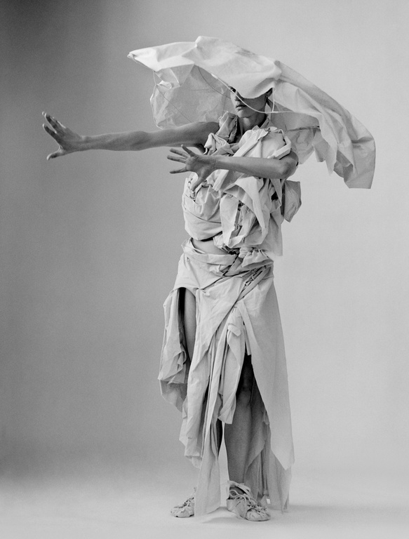 Black and white photograph of a person draped in white fabrics and paper, posing in a studio setting with outstretched arms, casting shadows.
