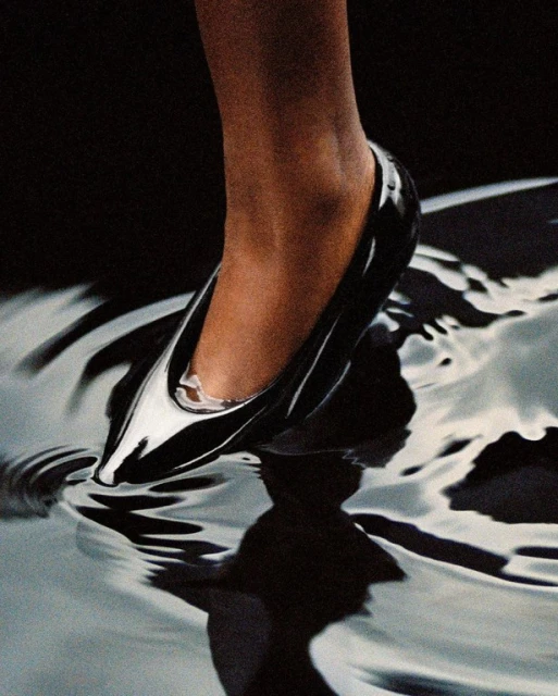 Close-up of a glossy black stiletto heel sinking into reflective rippled water, capturing dark elegance and fluid motion.