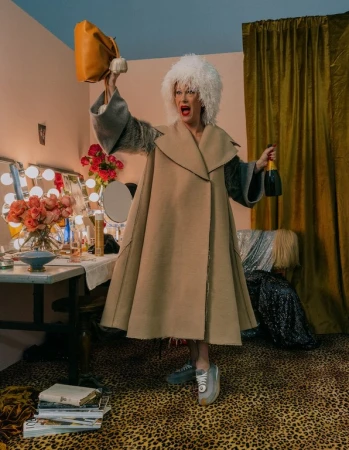 Woman in beige coat and white wig holds champagne and purse in a whimsical, colorful indoor setting with makeup table.