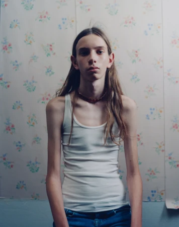 Young person with long brown hair wearing a white tank top and blue jeans against a vintage floral wallpaper background.
