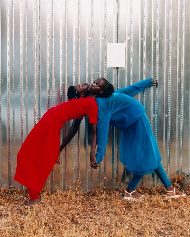 Two African women leaning back to back against a metal wall. One wears a red dress, the other a blue pantsuit. Neutral color palette.
