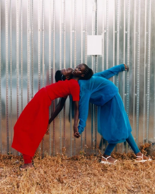 Two African women leaning back to back against a metal wall. One wears a red dress, the other a blue pantsuit. Neutral color palette.