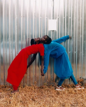 Two African women leaning back to back against a metal wall. One wears a red dress, the other a blue pantsuit. Neutral color palette.