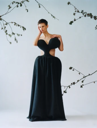 Model with short hair wearing a structured black sculptural dress against a plain background with hanging branches