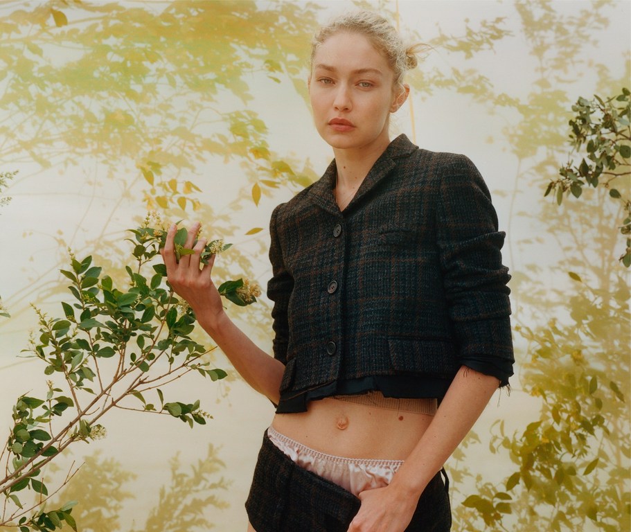 Model poses in a cropped tweed jacket and shorts, holding foliage against a neutral background with foliage shadows and diffuse light.