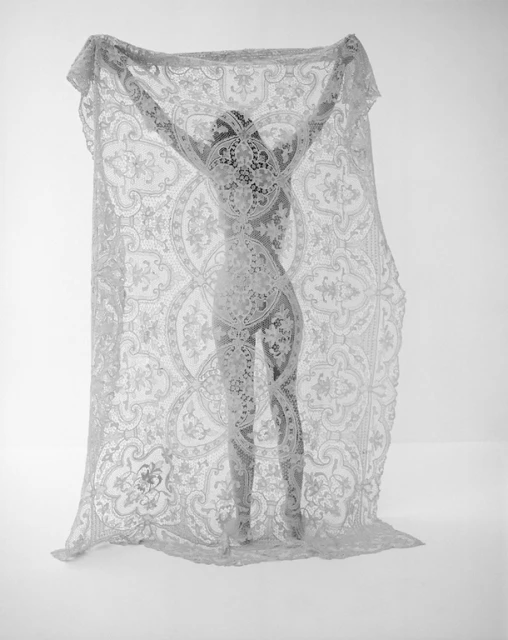 Monochrome studio shot of an adult woman posing behind a patterned lace curtain, creating a silhouette effect against a white backdrop.