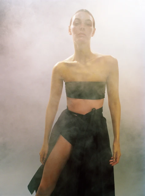 A woman in a strapless top and wrap skirt poses in a hazy studio setting with neutral colors. Fashion editorial shot.