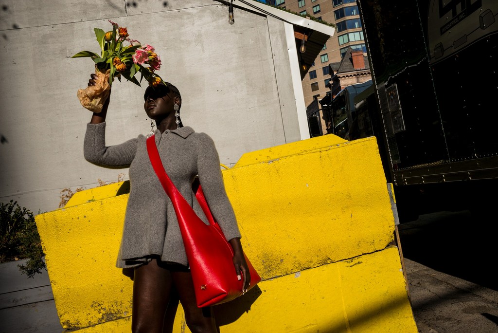 Woman in a grey coat poses with flowers and a red purse against a bright yellow wall in an urban setting on a sunny day.