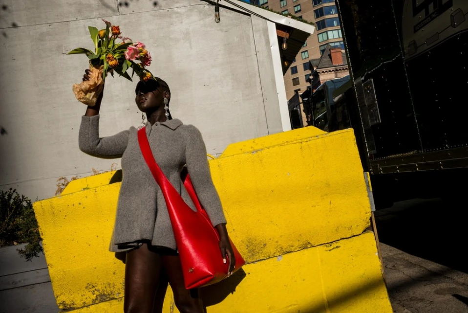 Woman in a grey coat poses with flowers and a red purse against a bright yellow wall in an urban setting on a sunny day.