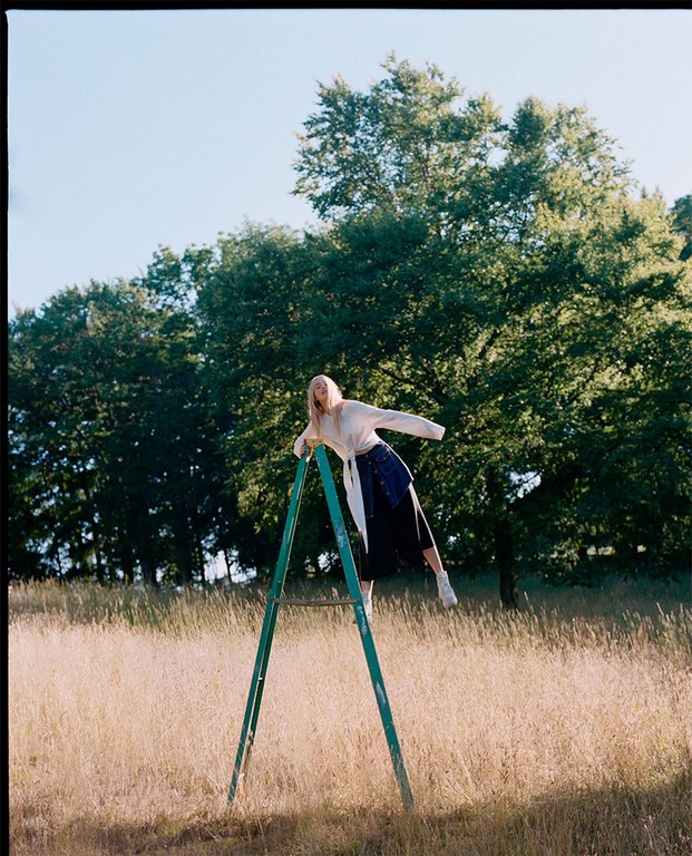 Woman in white sweater and skirt poses on metal frame in grassy field with trees under blue sky in fashion photography.
