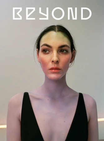 Portrait of a woman wearing a black top with green and pink makeup on her face against a plain light wall.
