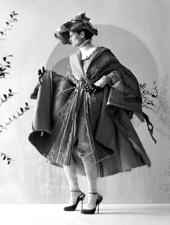 Model in wide-brimmed hat wearing a textured pleated coat and heels against a backdrop