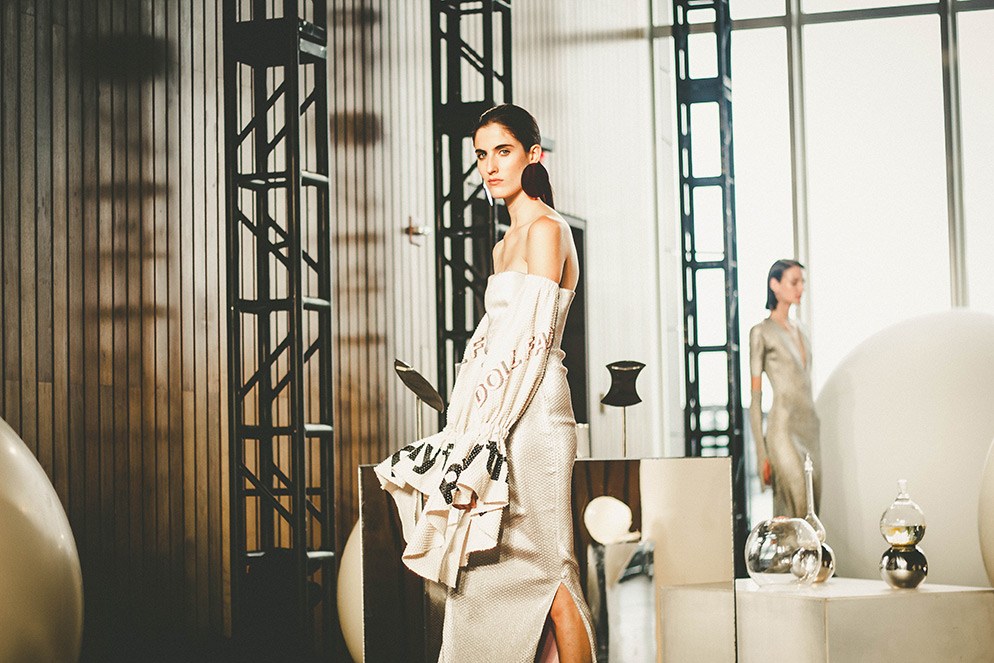 A woman in an off-the-shoulder dress with ruffled sleeves, standing in front of a wood panel and geometric metal structures inside.