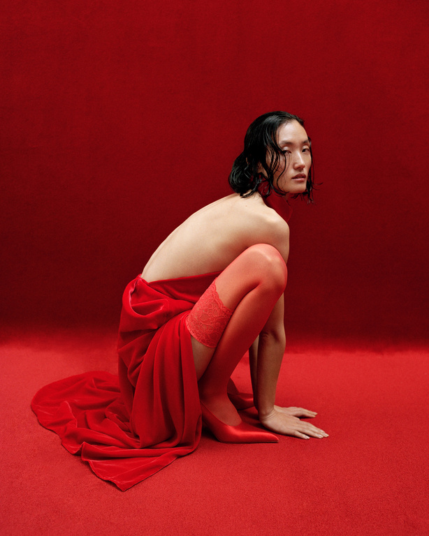 Fashion shot of an Asian adult female squatting with a red velvet drape over her body and wearing red hosiery and red pointed-toe heels.