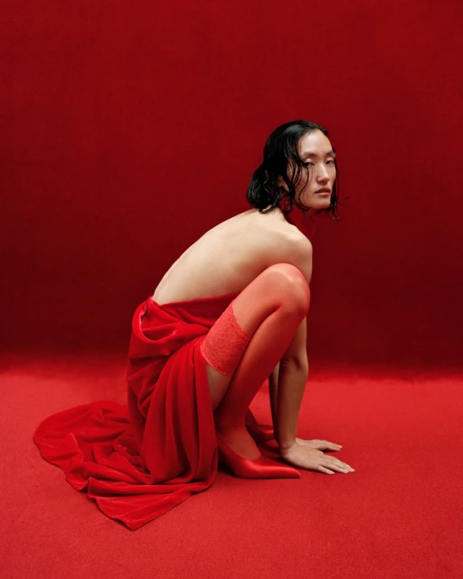Fashion shot of an Asian adult female squatting with a red velvet drape over her body and wearing red hosiery and red pointed-toe heels.