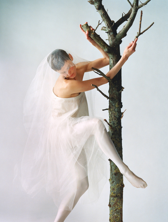 A woman with a veil and white clothing is climbing a bare tree against a plain white backdrop in a studio setting, portrait orientation.