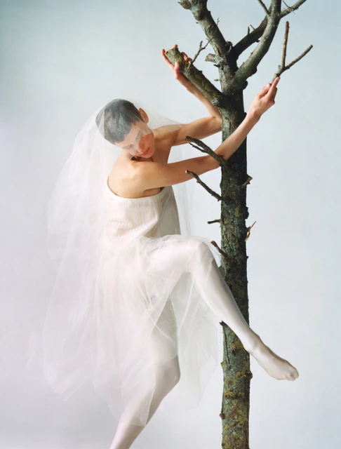 A woman with a veil and white clothing is climbing a bare tree against a plain white backdrop in a studio setting, portrait orientation.