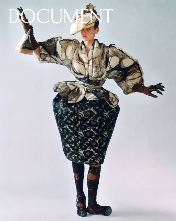 Model in avant-garde sculpted jacket and patterned skirt wearing a tilted hat against a white background