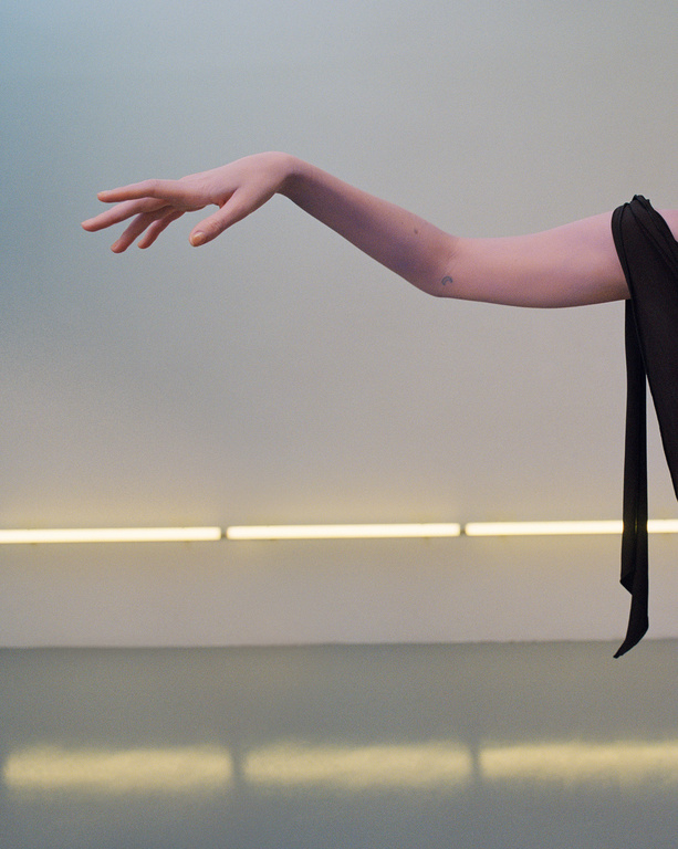 Elegant arm extends from black fabric against a pale wall with linear lighting, creating a minimalist and striking image.