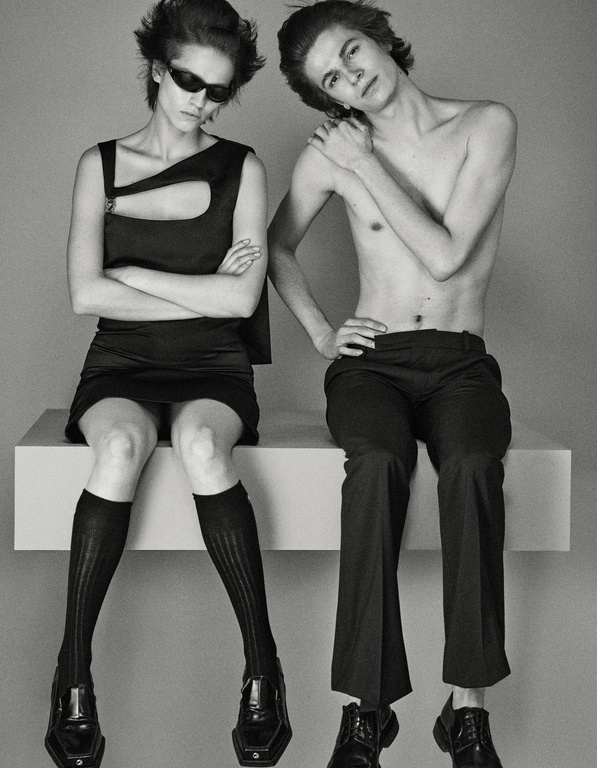 Two fashion models, a man and a woman, seated indoors in a studio against a gray backdrop. The woman wears sunglasses, a black dress, and black socks.