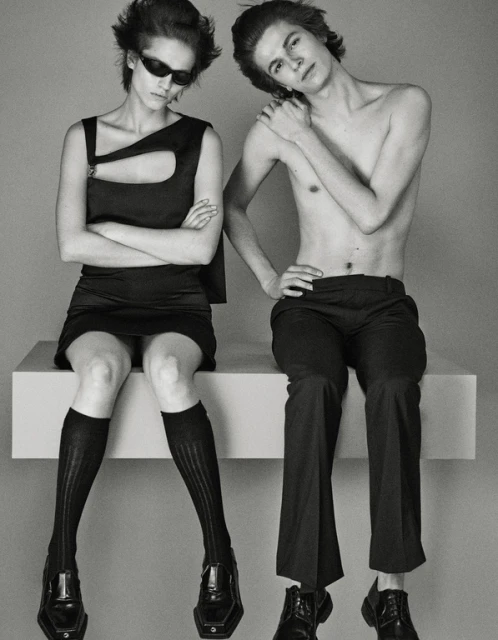 Two fashion models, a man and a woman, seated indoors in a studio against a gray backdrop. The woman wears sunglasses, a black dress, and black socks.