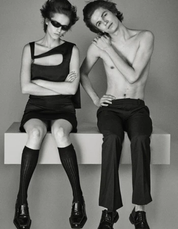 Two fashion models, a man and a woman, seated indoors in a studio against a gray backdrop. The woman wears sunglasses, a black dress, and black socks.