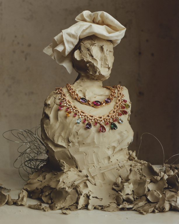 Clay sculpture of a bust with a fabric head covering and a gold chain necklace with colorful gemstones in a neutral studio setting.