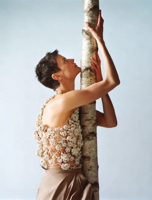 A woman embraces a birch tree, wearing a textured top and tan skirt against a light blue background, captured in a medium shot.
