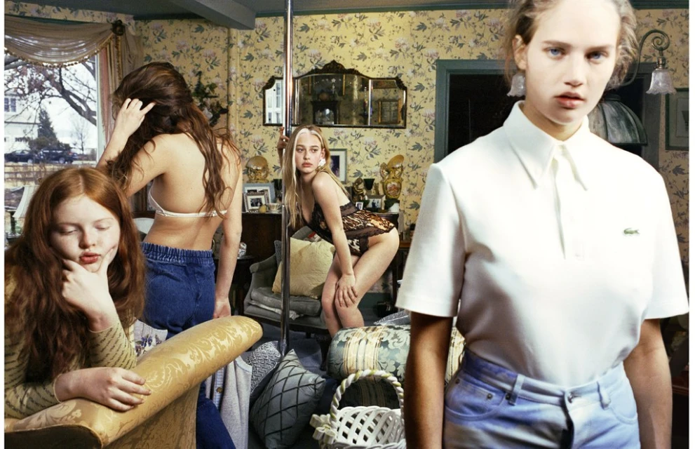 Four young women pose in a cluttered, vintage-style interior. One wears a white Lacoste polo shirt. Floral wallpaper backdrop with mirror.