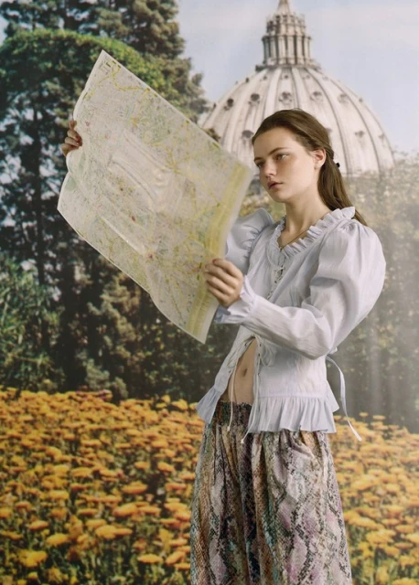 A young adult female stands in front of a scenic backdrop, holding a map. She is wearing a light blue top and patterned pants.