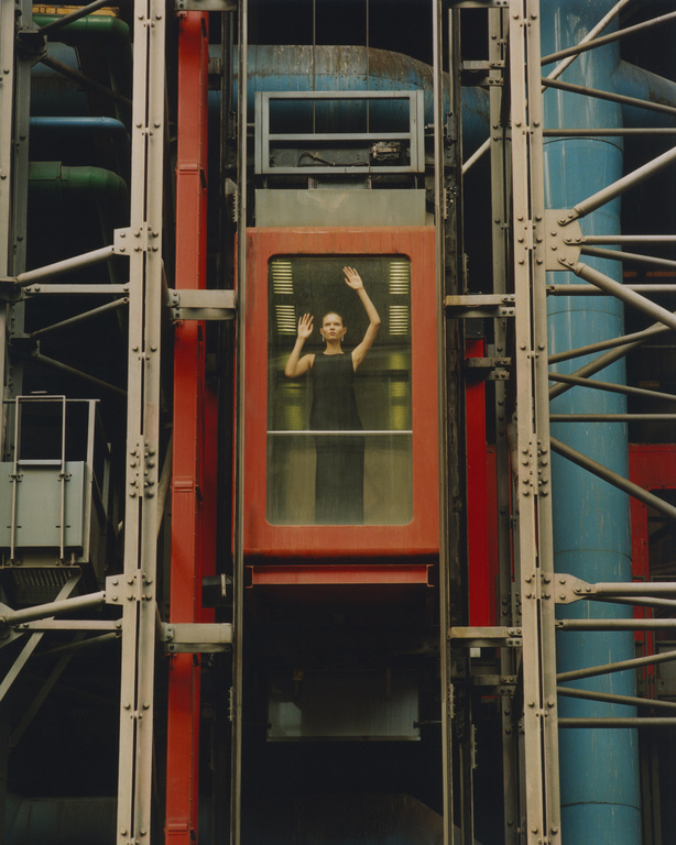 Woman stands inside a red elevator with hands raised, framed by industrial architecture with blue and green pipes and metal beams.
