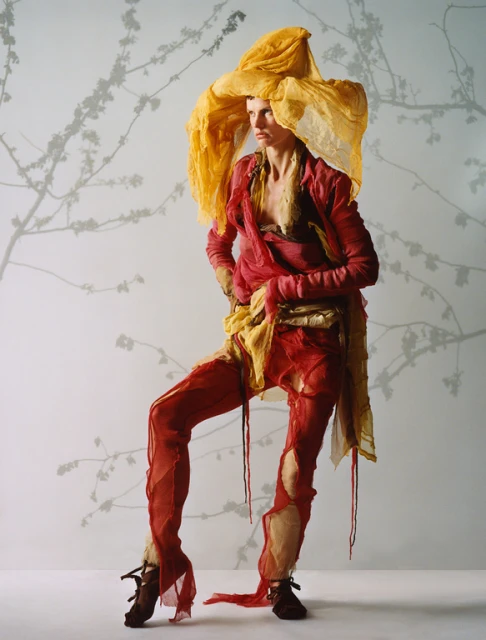 Woman in Avant-Garde Fashion: Voluminous yellow hat, red top and pants, textured fabrics. Studio portrait with floral-branch backdrop.