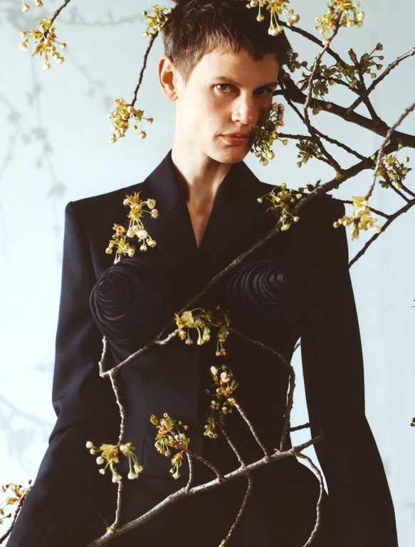 Close-up portrait of a fair-skinned adult in a stylish black jacket, surrounded by blossoming branches against a soft light blue backdrop.