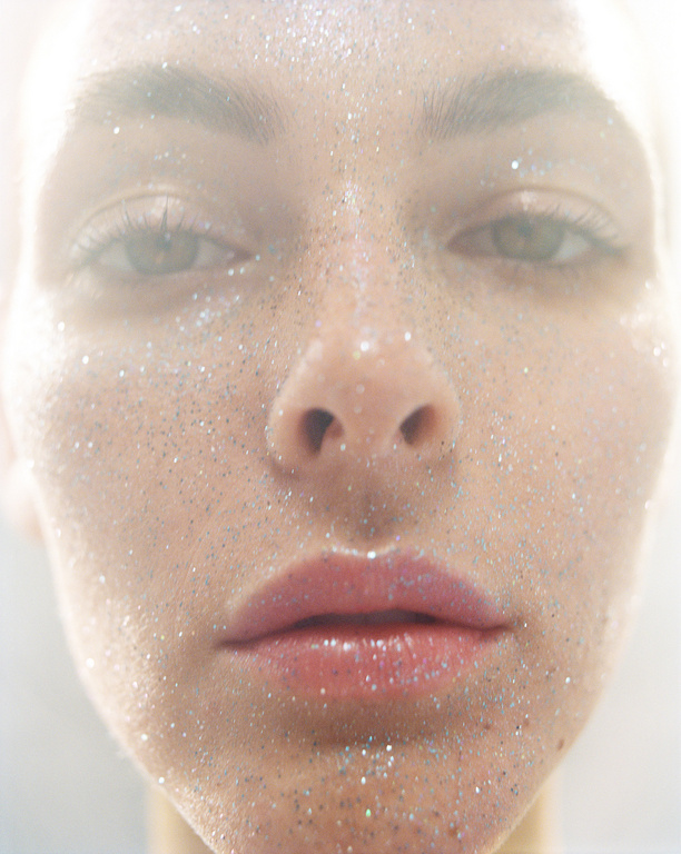 Close-up portrait of a young adult woman with glitter on her face, fair skin, and light eye color, set against a bright, hazy background.