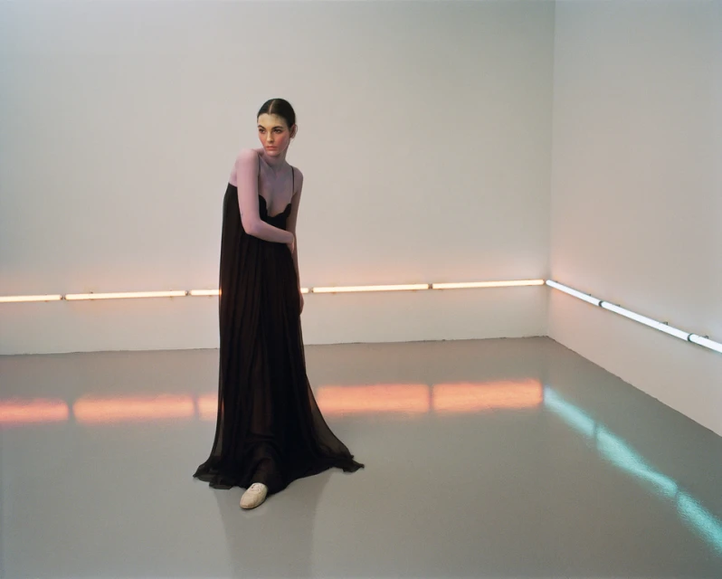 Woman in a long black dress posing in a photo studio with neon lighting in the background. Minimalist, full-length shot in a sparse setting.
