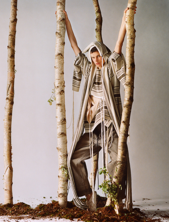 Fashion shot of an androgynous model wearing a hooded coat and pants posing between birch trees in a studio setting.