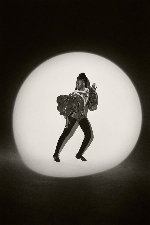 Woman in sequined top and black leggings strikes a pose in a spotlight against a dark backdrop, showcasing fashion and dance.